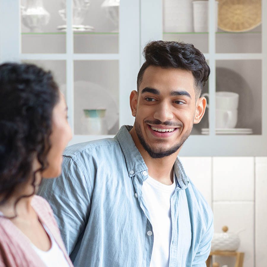 A young man with dark hair and a beard, wearing a light blue shirt, smiles and talks to a woman in a kitchen with glass-front cabinets in the background.