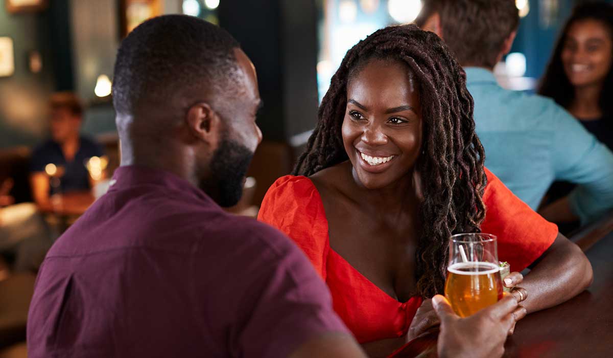 A woman in an orange top smiles while talking to a man at a bar. She holds a glass of beer, and both appear to be enjoying their conversation in a relaxed, dimly lit setting. Other people are visible in the background.