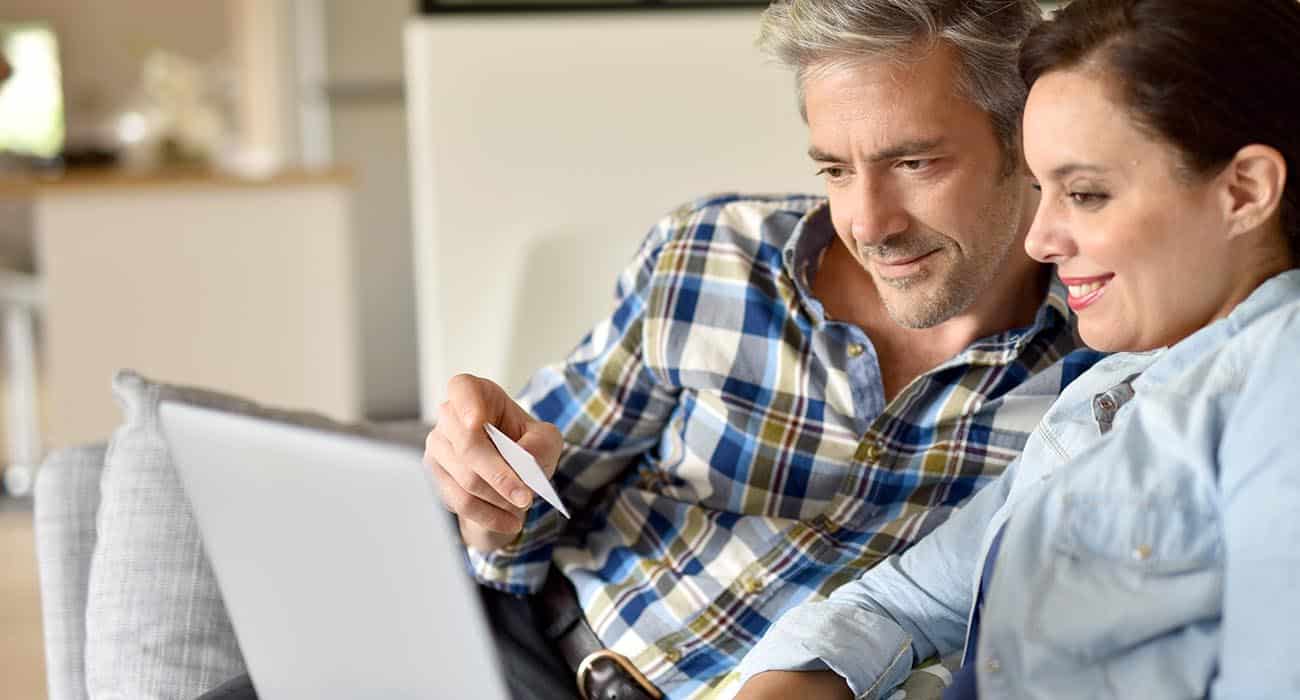 Three young women wearing sunglasses and sun hats excitedly look into shopping bags outdoors on a sunny day, smiling and appearing pleasantly surprised by their purchases.