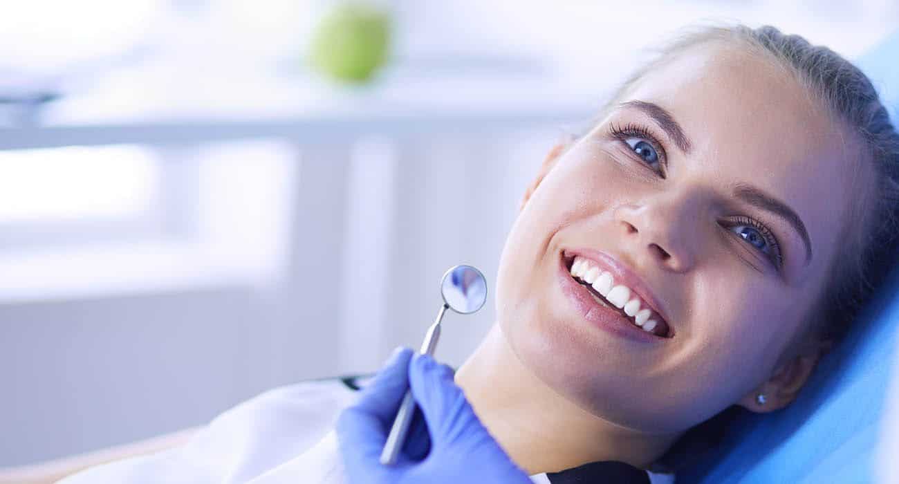 A young woman smiles and reclines in a dental chair while a dentist wearing blue gloves holds a dental mirror near her mouth during an examination.