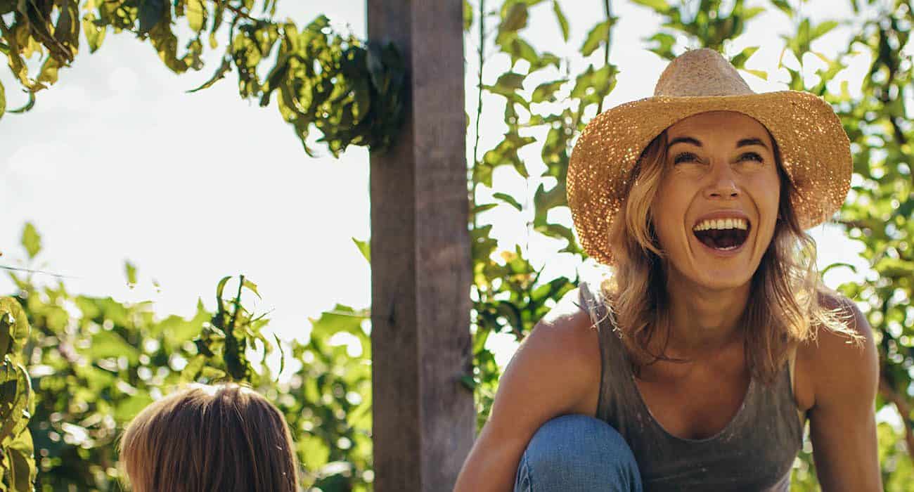 A woman wearing a straw hat and a sleeveless top laughs joyfully outdoors, surrounded by green leafy plants and sunlight.