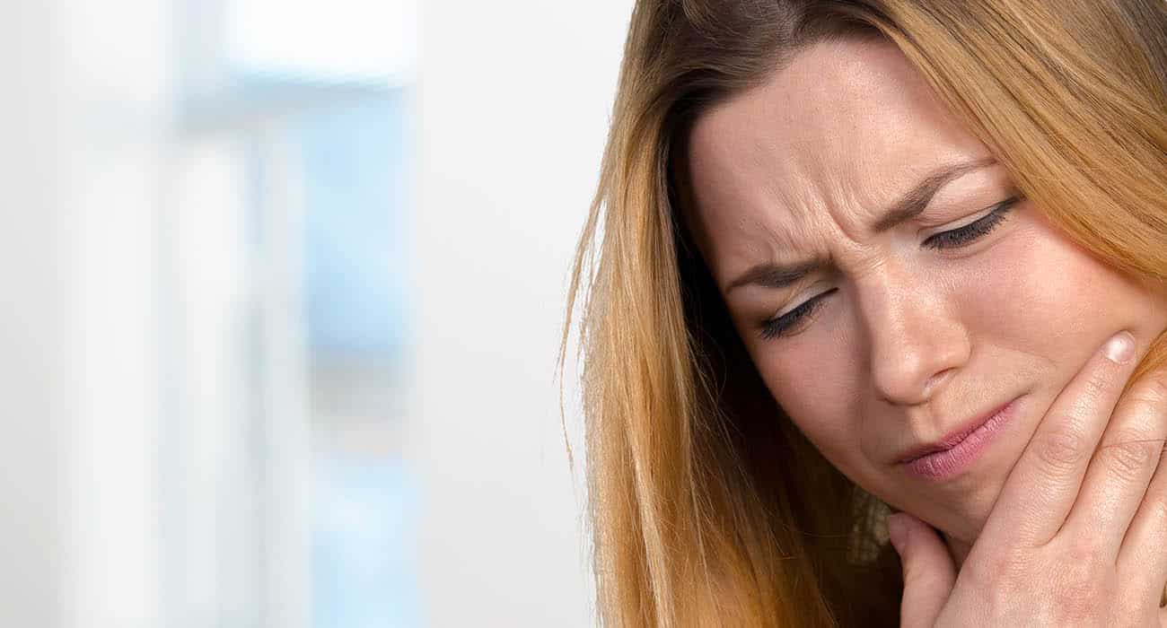A woman sits indoors with her eyes closed, holding her cheek and wincing in pain, suggesting she has a toothache or facial discomfort.