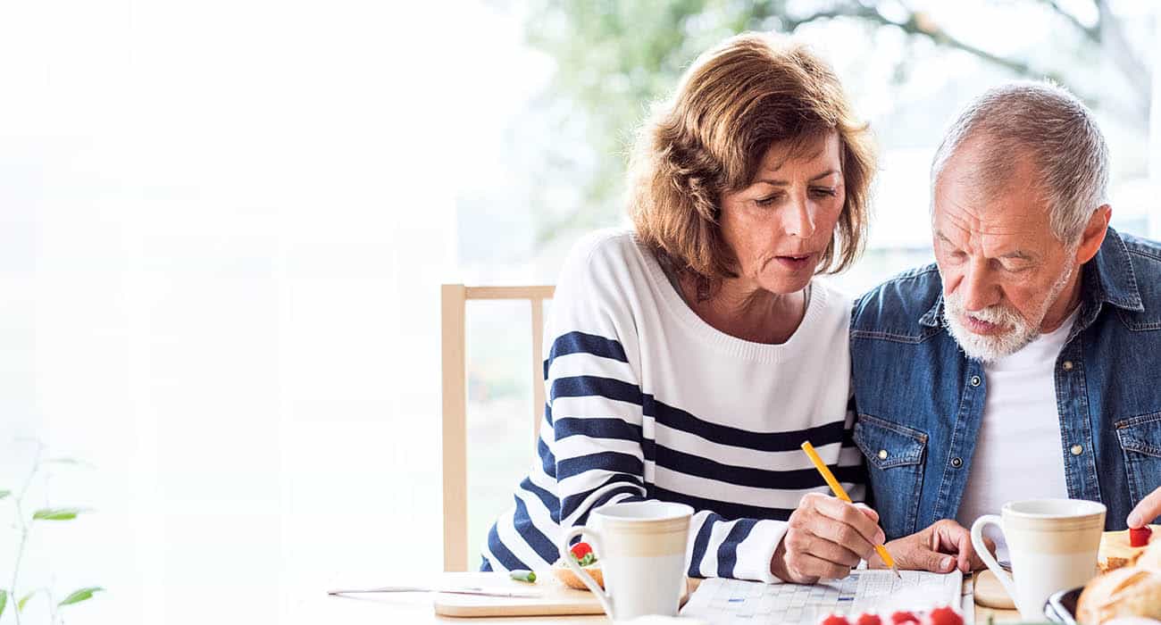 A man with short dark hair and a beard smiles while looking slightly to the side, wearing a green sweater. The background is a brightly lit, blurred indoor setting.