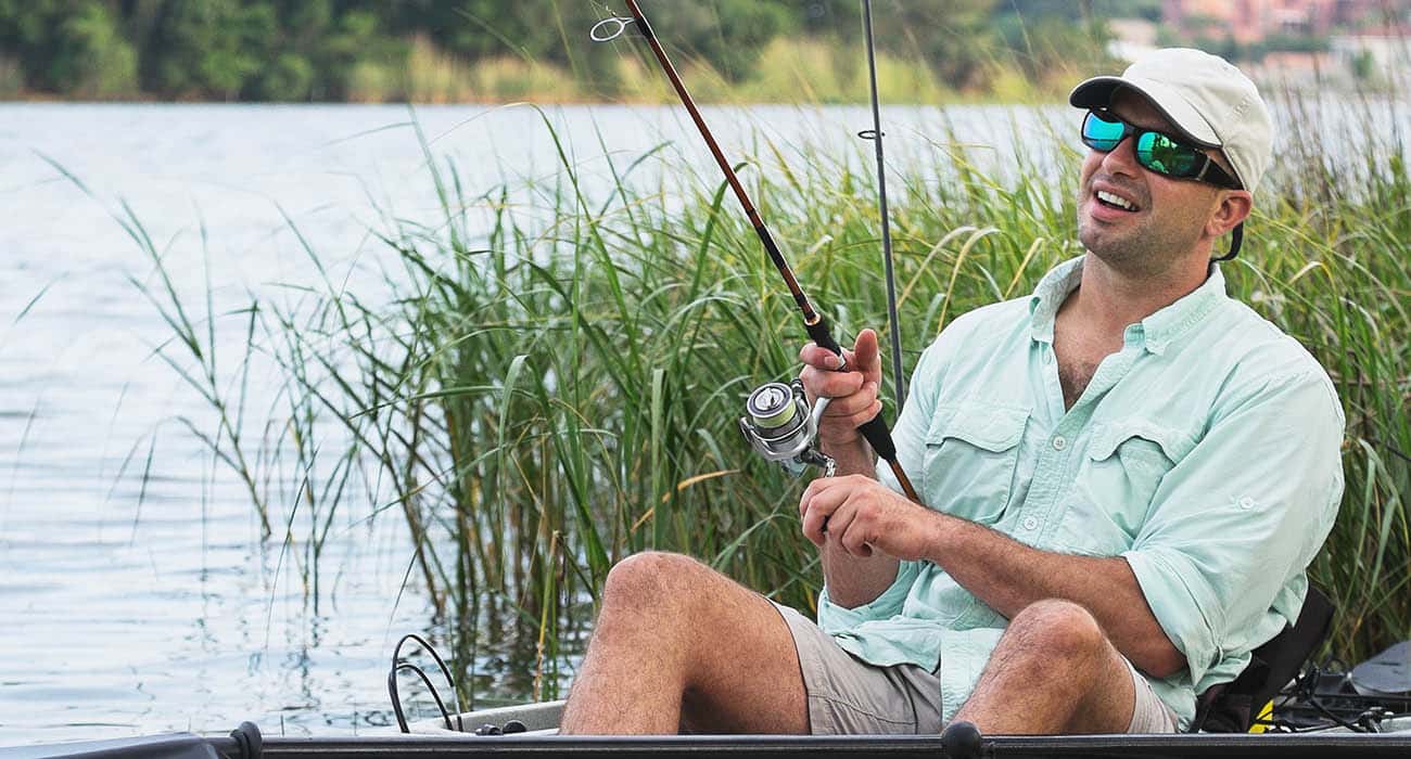 A man wearing sunglasses, a light-colored cap, and a light shirt smiles while fishing from a small boat surrounded by reeds on a lake.