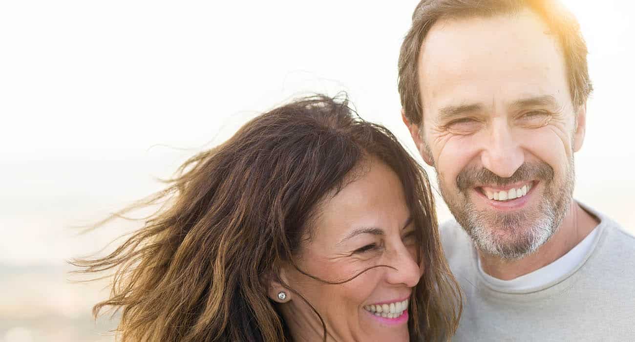 A woman with medium-length dark hair and a white shirt smiling warmly at the camera, with a bright, softly blurred background.
