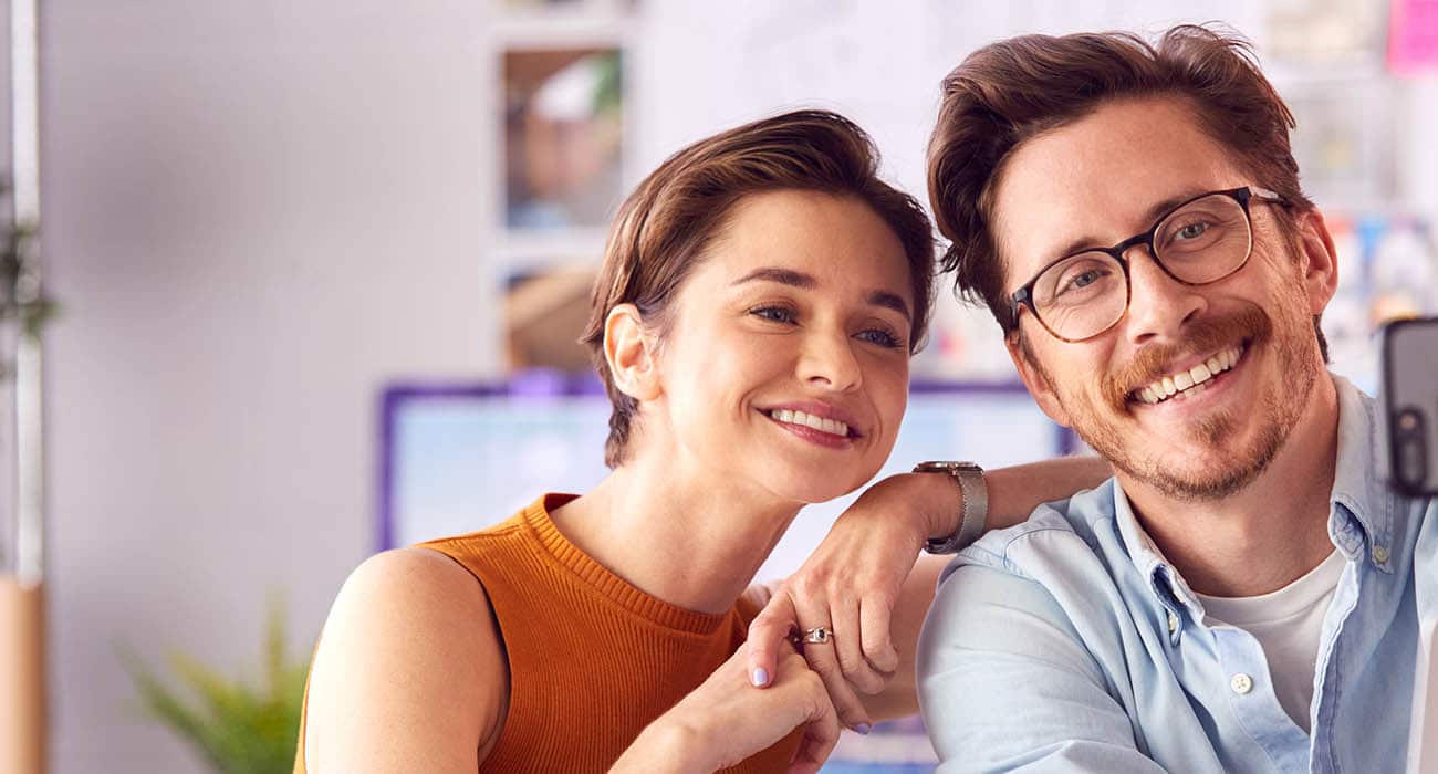 A smiling woman in an orange sleeveless top and a man with glasses in a light blue shirt pose closely together, taking a selfie indoors.