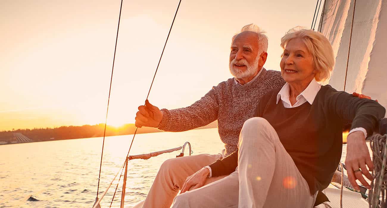 An elderly couple sits together on a sailboat at sunset, smiling and relaxed, with warm sunlight illuminating their faces and the sail in the background.